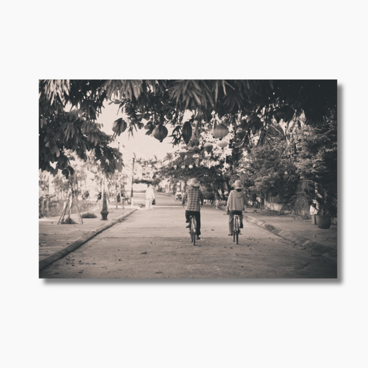 Couple cycling down a tree-lined street in Hoi An, Vietnam — limited edition fine art travel and romance photography print.
